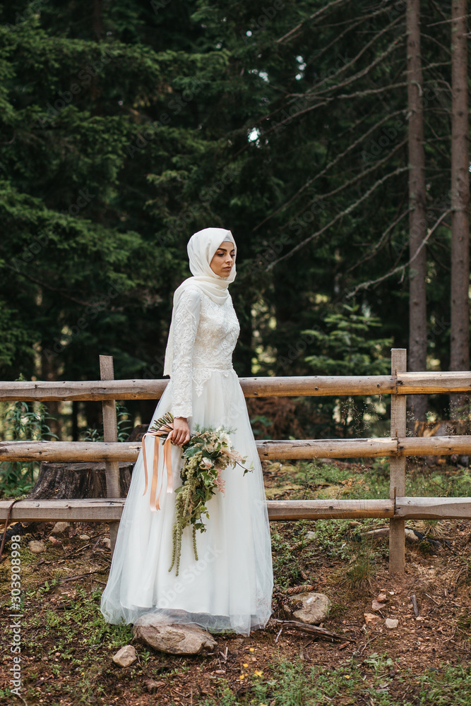 Beautiful elegant bride with hijab in lace wedding dress with long full skirt and long sleeves. She is holding a big bouquet of flowers. Outdoors, in the woods.