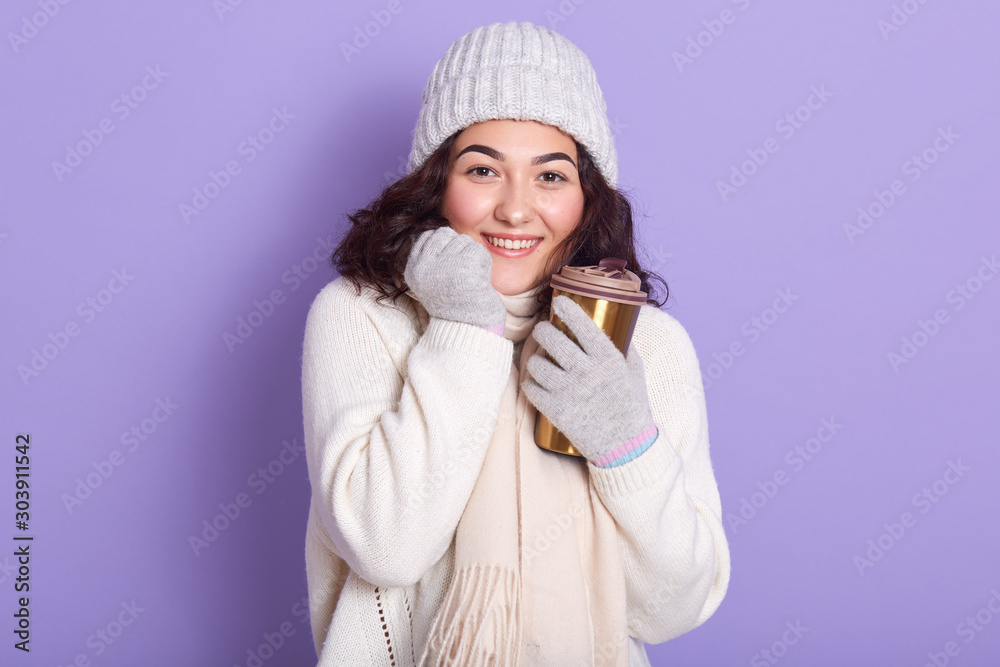 Young attarctive woman basking her hands via cup of hot beverage and gloves, wearing sweater, scarf and cap, posing isolated over purple background, using thermo mug, moving towards zero waste. Young attarctive woman basking her hands via cup of hot beverage and gloves, wearing sweater, scarf and cap, posing isolated over purple background, using thermo mug, moving towards zero waste.