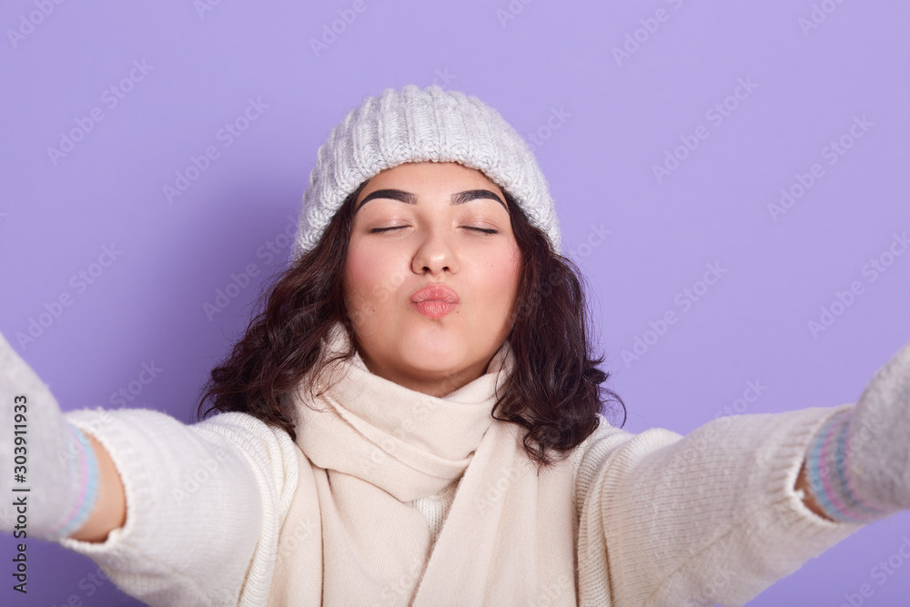 Woman making kiss gesture towards camera and spreading hands aside, keeping eyes closed, female with dark hair wearing white warm sweater, cap and scarf, posing isplated over lilac studio background. Woman making kiss gesture towards camera and spreading hands aside, keeping eyes closed, female with dark hair wearing white warm sweater, cap and scarf, posing isplated over lilac studio background.