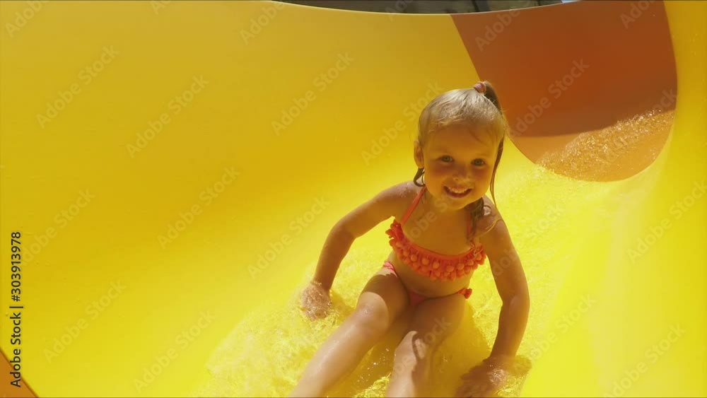 Funny little girl rides down an orange slide in a water park. Girl in a swimsuit laughs and slides into the water raising spray