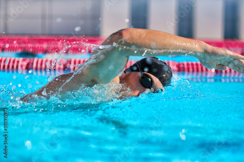 Swimmer man doing crawl swim in swimming pool portrait. Closeup of athlete wearing goggles, swimming cap training in blue water indoors.