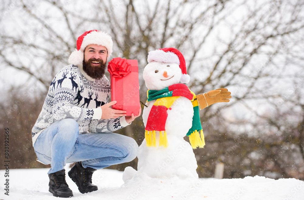 Winter vacation. Man made snowman. Man Santa hat having fun outdoors. Winter games. Guy happy face snowy nature background. Hipster with beard hold gift box. Surprise concept. Winter activity