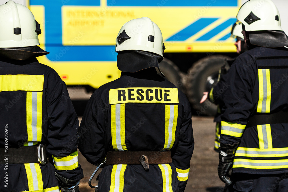 Group of firefighters in safe helmet and uniform standing by car Group of firefighters in safe helmet and uniform standing by car