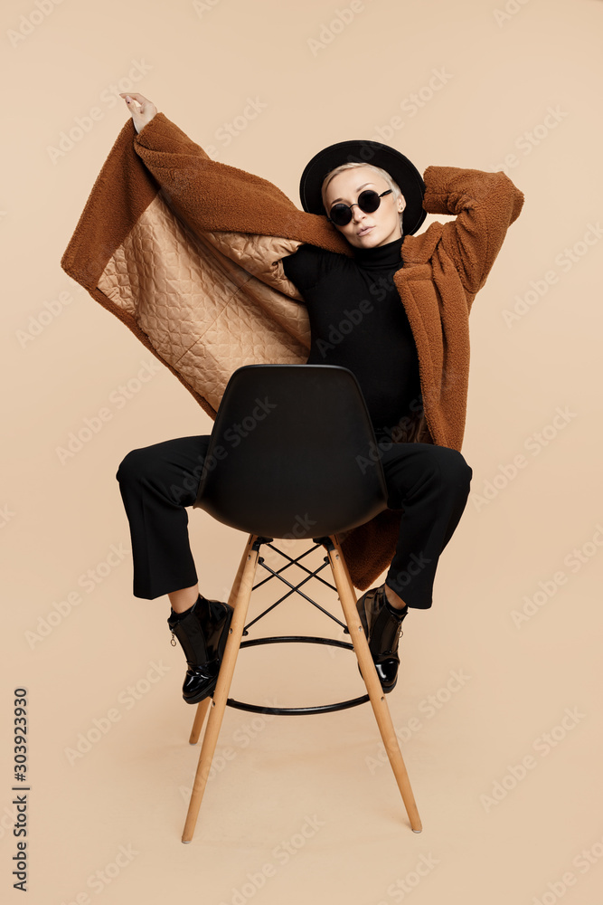 Φωτογραφία Trendy young hipster woman with blonde short hair wearing a coat, hat and sunglasses posing over beige background