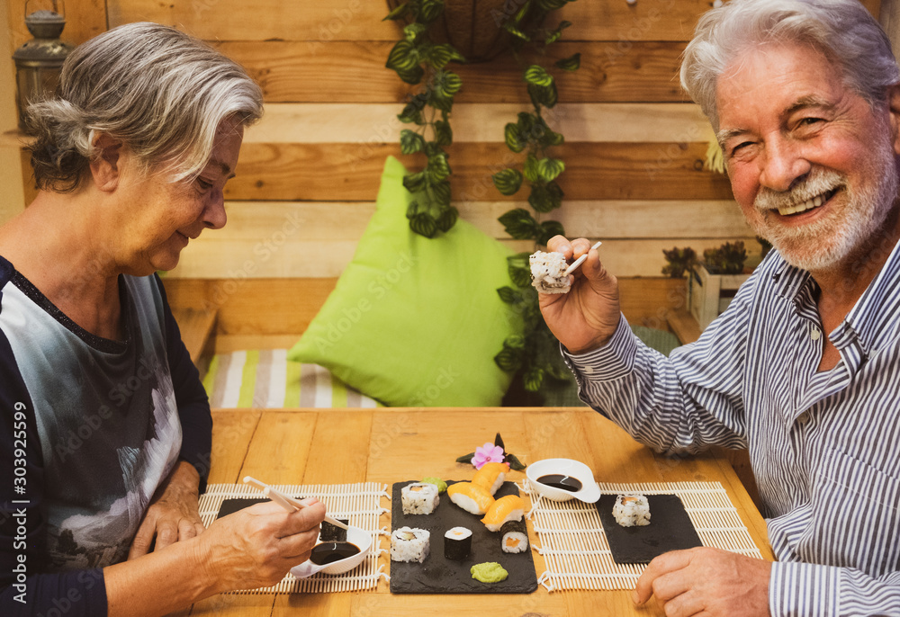 Cheerful elderly man with white hair and beard enjoying japanese food with his wife. Outdoors on a wooden table with sushi on the table Cheerful elderly man with white hair and beard enjoying japanese food with his wife. Outdoors on a wooden table with sushi on the table