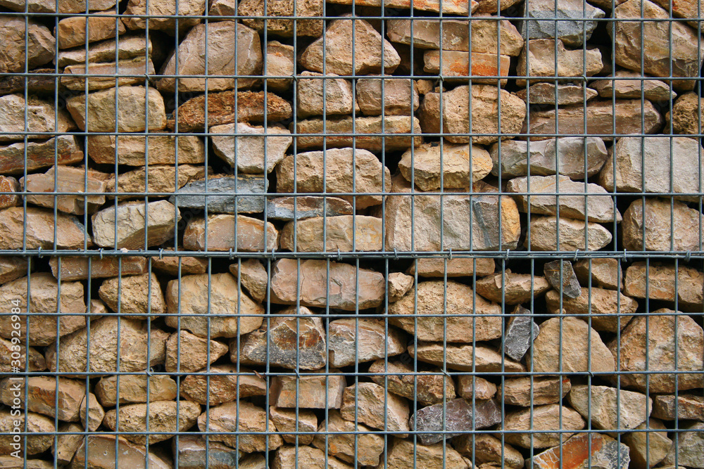 Stone wall of grindstone of light and gray color enclosed behind a metal grid. Mineral surface without mortar joints. Construction for old house and park. Stone wall of grindstone of light and gray color enclosed behind a metal grid. Mineral surface without mortar joints. Construction for old house and park.