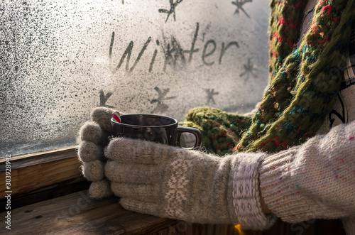 mug with hot drink coffee or tea in the hands of a girl in a sweater and mittens on the background of a frozen winter window, winter mood