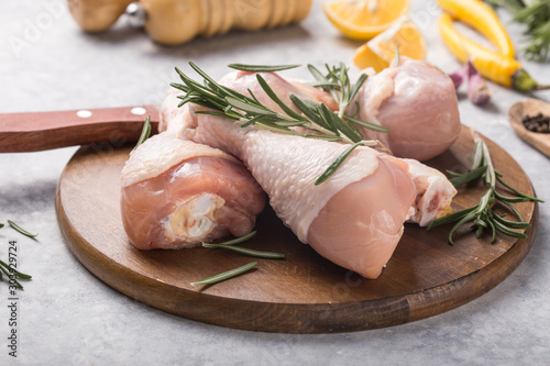 Raw organic uncooked chicken legs or drumsticks on round wooden board with ingredients for cooking (pepper, lemon, rosemary, salt, garlic). Meat top view