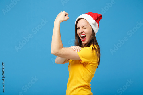 Side view of young woman Santa girl in yellow t-shirt Christmas hat posing isolated on blue background. New Year 2020 celebration holiday concept. Mock up copy space. Showing biceps, muscles blinking. © ViDi Studio