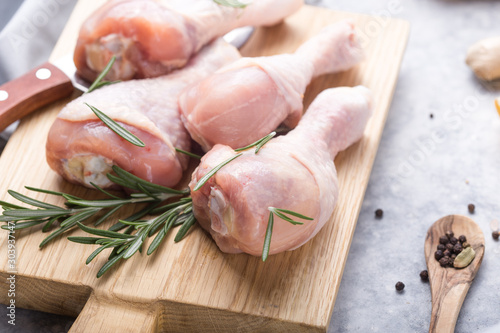 Raw organic uncooked chicken legs or drumsticks on cutting board with ingredients for cooking (pepper, lemon, rosemary, salt, garlic). Meat top view