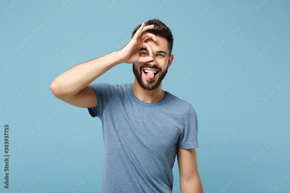 Young cheerful man in casual clothes posing isolated on blue wall background. People lifestyle concept. Mock up copy space. Showing tongue, holding hands near eyes, imitating glasses or binoculars. Young cheerful man in casual clothes posing isolated on blue wall background. People lifestyle concept. Mock up copy space. Showing tongue, holding hands near eyes, imitating glasses or binoculars.