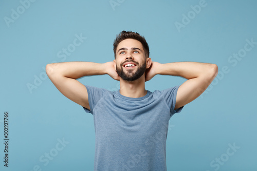 Young smiling man in casual clothes posing isolated on blue wall background, studio portrait. People sincere emotions lifestyle concept. Mock up copy space. Looking up with hands behind his head. © ViDi Studio