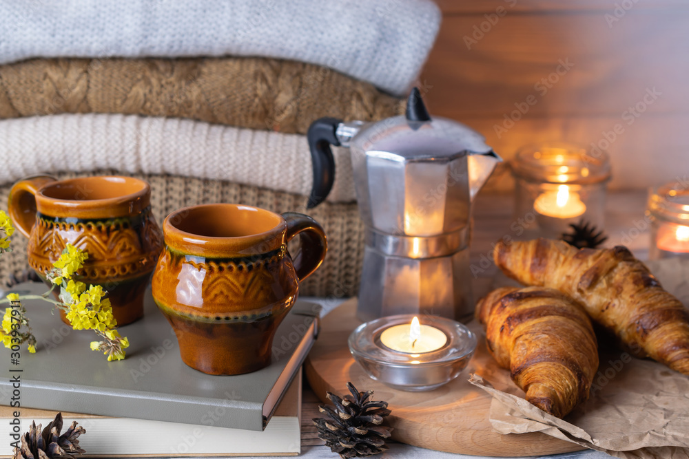 Hygge composition with two rustic clay cups, croissants, books, candles and knitwear on wooden table