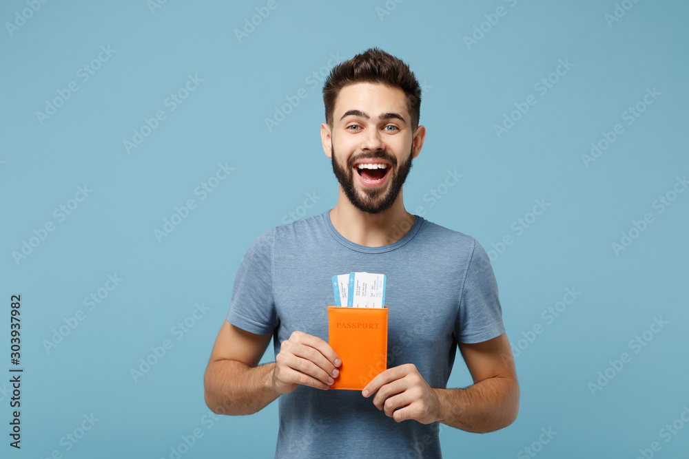 Young laughing funny man in casual clothes posing isolated on blue wall background, studio portrait. People lifestyle concept. Mock up copy space. Holding in hands passport, boarding pass tickets.