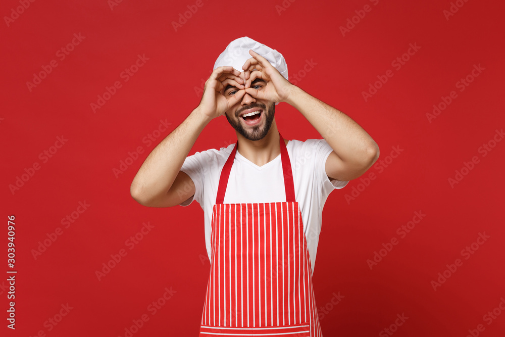 Bearded male chef cook or baker man in striped apron toque chefs hat posing isolated on red background. Cooking food concept. Mock up copy space. Hold hands near eyes, imitating glasses or binoculars. Bearded male chef cook or baker man in striped apron toque chefs hat posing isolated on red background. Cooking food concept. Mock up copy space. Hold hands near eyes, imitating glasses or binoculars.