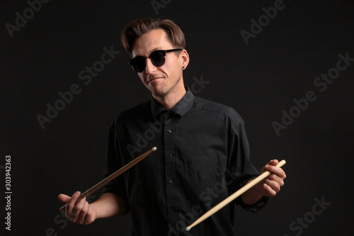 Stylish man drummer in a black shirt and sun glasses playing drums with sticks over dark background.