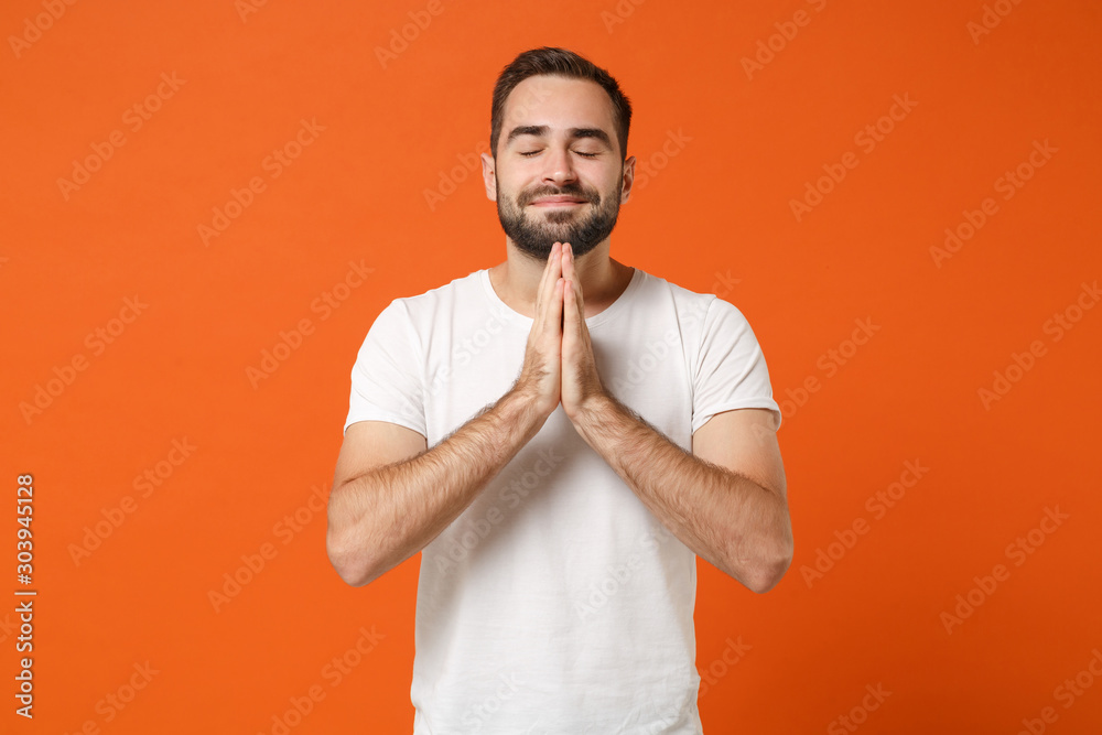 Young man in casual white t-shirt posing isolated on bright orange wall background, studio portrait. People lifestyle concept. Mock up copy space. Holding hands folded in prayer, keeping eyes closed. Young man in casual white t-shirt posing isolated on bright orange wall background, studio portrait. People lifestyle concept. Mock up copy space. Holding hands folded in prayer, keeping eyes closed.