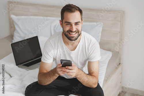 Young bearded man sits in bed with white sheet pillow blanket in bedroom at home. Male spending time in room using mobile phone, display pc. Rest relax good mood lifestyle concept. Mock up copy space.