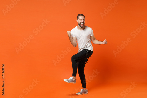 Joyful happy young man in casual white t-shirt posing isolated on orange wall background studio portrait. People sincere emotions lifestyle concept. Mock up copy space. Clenching fists like winner.