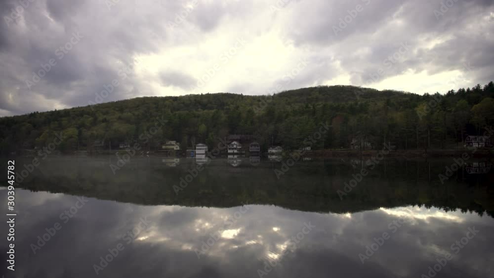 Clouds float over a glassy still lake reflecting tthe surrounding houses and forrest covering the hills. Clouds float over a glassy still lake reflecting tthe surrounding houses and forrest covering the hills.