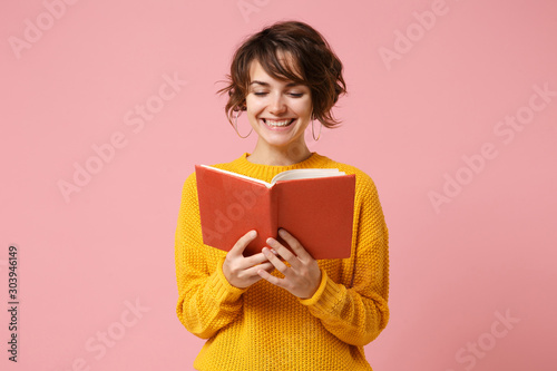 Smiling young brunette woman girl in yellow sweater posing isolated on pink wall background, studio portrait. People sincere emotions lifestyle concept. Mock up copy space. Holding and reading book.
