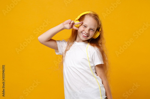 Little ginger kid girl 12-13 years old in white t-shirt isolated on yellow background studio portrait. Childhood lifestyle concept. Mock up copy space. Listen music in headphones showing victory sign.