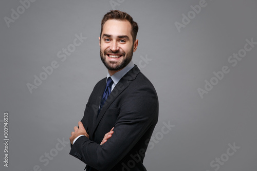 Side view of young business man in classic black suit shirt tie posing isolated on grey background. Achievement career wealth business concept. Mock up copy space. Hold hands crossed, looking camera.
