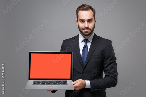 Handsome young business man in black suit shirt tie posing isolated on grey background. Achievement career wealth business concept. Mock up copy space. Hold laptop pc computer with blank empty screen.