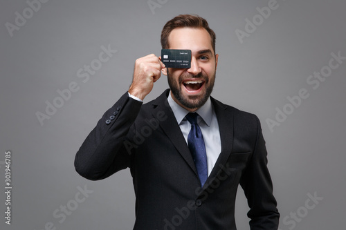 Cheerful young business man in classic black suit shirt tie posing isolated on grey wall background. Achievement career wealth business concept. Mock up copy space. Covering eye with credit bank card.
