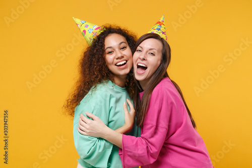 Two cheerful women friends european and african american girls in pink green clothes, birthday hats posing isolated on yellow orange background. People lifestyle concept. Mock up copy space. Hugging.