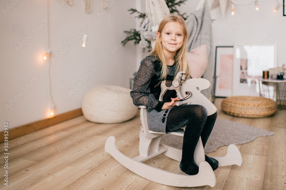 Φωτογραφία Little happy blonde girl riding toy horse in studio with cristmas tree and new year decorations