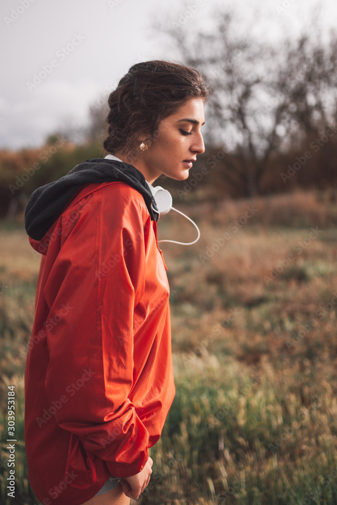 Naklejka premium Young woman standing with raincoat and headphones on the field