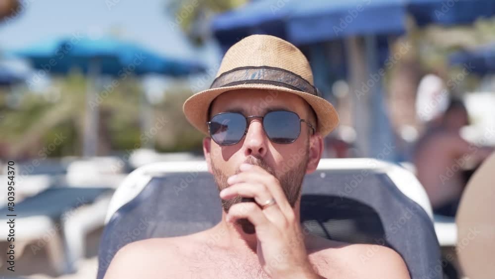 Young handsome man in a sunglasses is sitting on a sunbed on a beach and smoking an i-qos. Young handsome man in a sunglasses is sitting on a sunbed on a beach and smoking an i-qos.