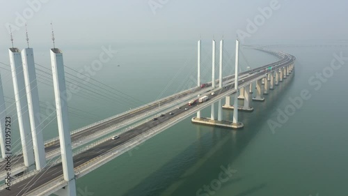 Retreating aerial footage of commuting traffic (passenger vehicles and cargo trucks) driving over massive Jiaozou Bay Bridge, connecting the more industrial Huangdao to residential Qingdao photo