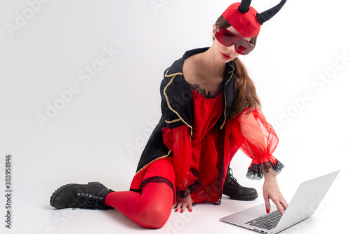 Sexy brunette woman in red tights, black boots and devil horns using laptop over white studio background. photo