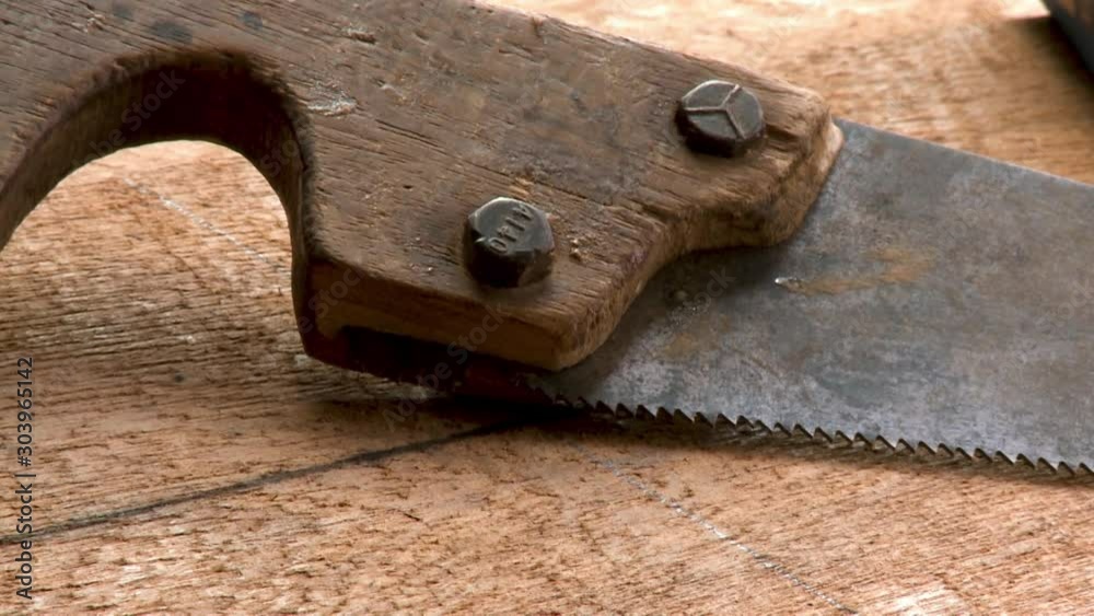 A daylight extreme closeup shot of an old saw with a rusty blade screwed on a wooden handle laying on top of a piece of wood inside a local woodworking shop.. A daylight extreme closeup shot of an old saw with a rusty blade screwed on a wooden handle laying on top of a piece of wood inside a local woodworking shop..