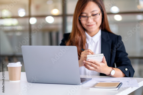 Closeup image of businesswoman writing on notebook while working on laptop in office