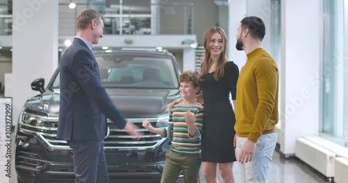 Caucasian car dealer standing in front of new car and congradulating young family with the purchase of automobile. Man shaking father's hand and giving high five to son. Cinema 4k footage ProRes HQ. photo