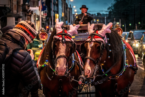 Horse wearing a hat with reindeer antlers and other Christmas decorations, haulin a cart decorated with christmas lights of multiple colors.