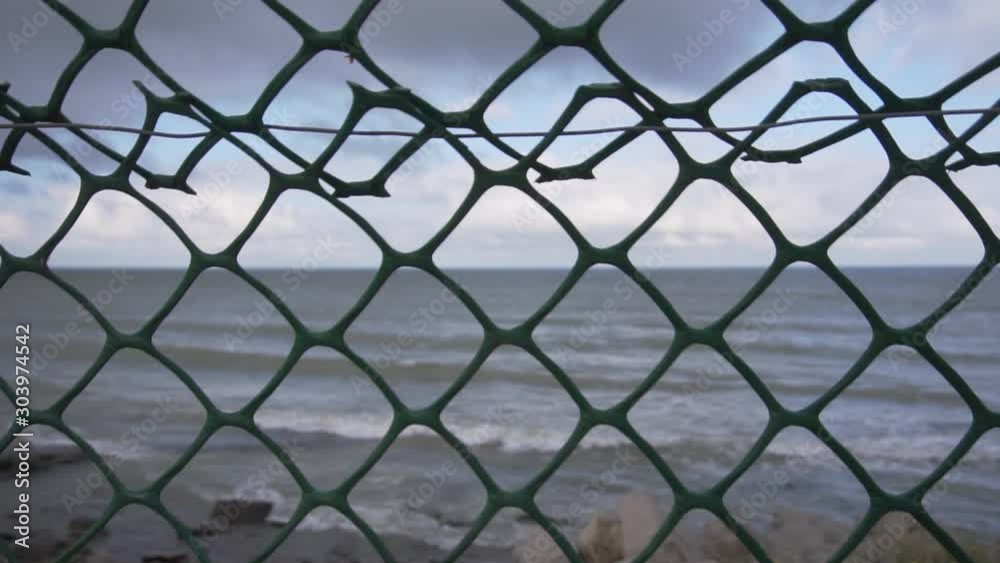 View through metallic net on beautiful coastline with big waves slowlyh falling on shores with stones and rocks on windy summer day. View through metallic net on beautiful coastline with big waves slowlyh falling on shores with stones and rocks on windy summer day.