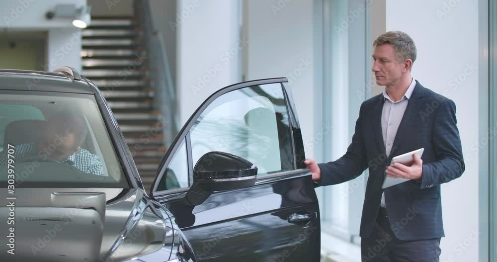 Confident Caucasian male trader with tablet opening car door for the customer in checkered shirt. Successful man choosing new automobile. Car dealership, car business. Cinema 4k footage ProRes HQ.