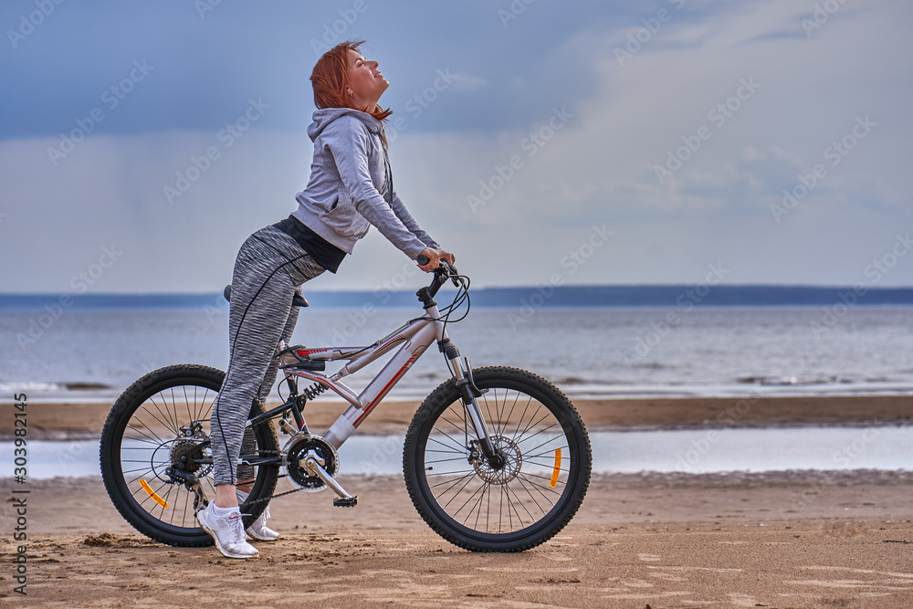 Red-haired middle-aged woman in sportswear on a bike walk on the sandy shore of a large river. Cloudy summer day.