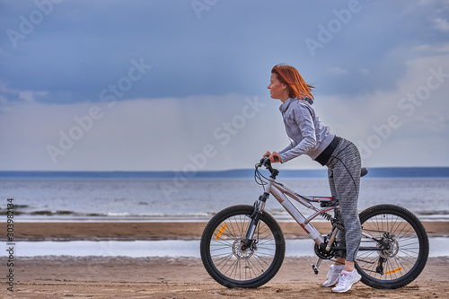 Red-haired middle-aged woman in sportswear on a bike walk on the sandy shore of a large river. Cloudy summer day.