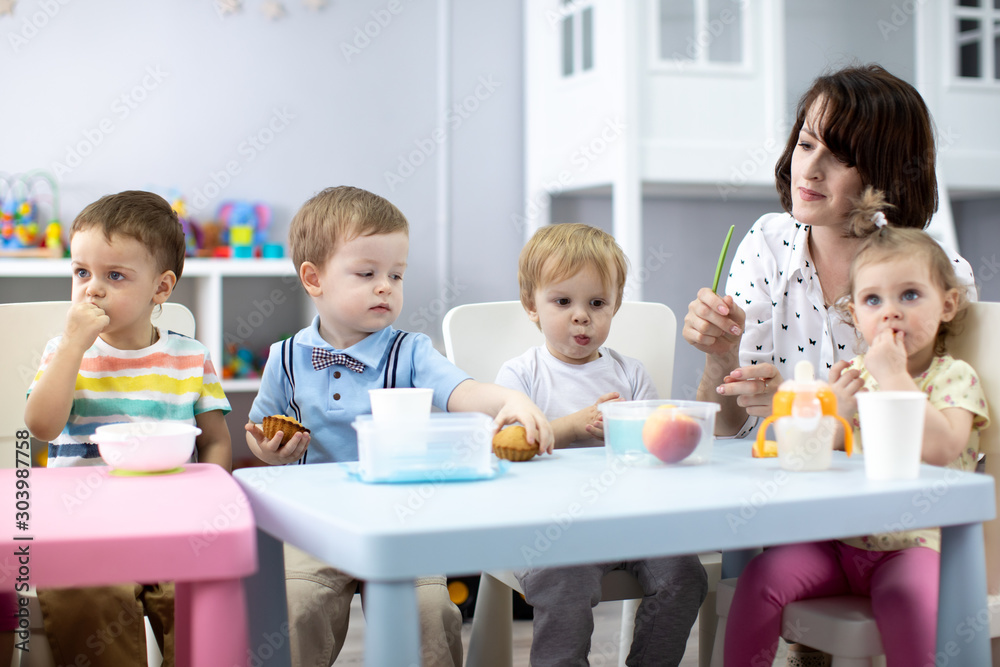 Kids group have lunch with teacher in nursery. Children eat food from lunch boxes in kindergarten Kids group have lunch with teacher in nursery. Children eat food from lunch boxes in kindergarten