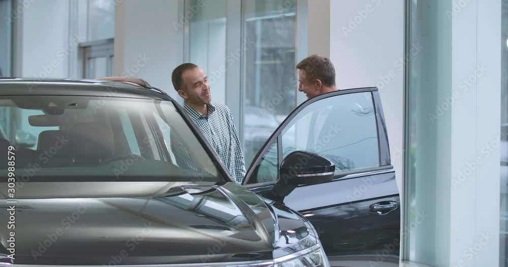 Two mature Caucasian men standing next to black automobile and shaking hands. Male car dealer making deal with the customer in auto showroom. Car dealership, car business. Cinema 4k footage ProRes HQ. Two mature Caucasian men standing next to black automobile and shaking hands. Male car dealer making deal with the customer in auto showroom. Car dealership, car business. Cinema 4k footage ProRes HQ.