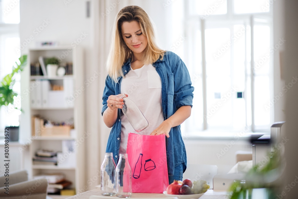 Young woman putting glass bottles in bag indoors at home, recycling concept. Young woman putting glass bottles in bag indoors at home, recycling concept.