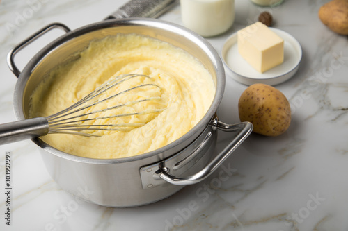 Kochtopf mit Kartoffelpüree mit Zutaten aus Kartoffeln, Milch, Salz, Butter, Muskatnuss dazu Muskatreibe und Schneebesen auf hell Marmor Hintergrund photo