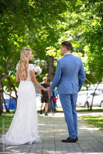 Young beautiful couple of newlyweds bride and groom walk holding hands in a park with green trees on their wedding day. Love and lovestory of wife and husband before starting a family.
