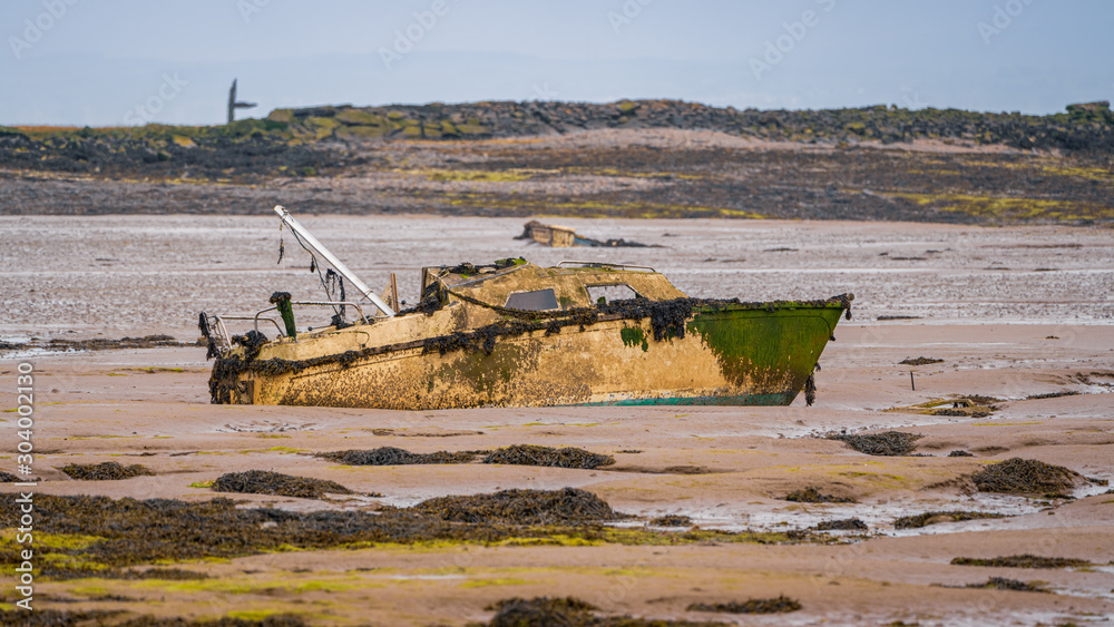 Fototapeta premium A damaged boat in the mud of the Walney Channel, seen from the road to Roa Island, Cumbria, England, UK