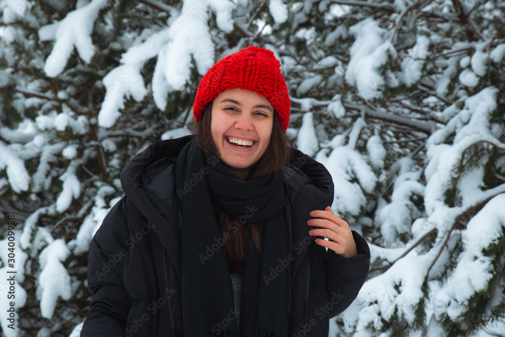 portrait of young smiling woman in winter clothes in red hat with bubo fir tree in background portrait of young smiling woman in winter clothes in red hat with bubo fir tree in background